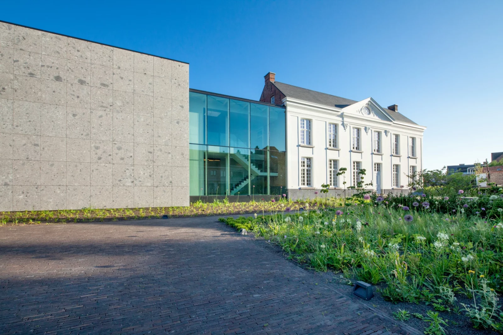 Het kantoorgebouw van project G01: een gerenoveerde witte pastorij met een glazen tussenstuk en een uitbreiding in natuursteen, met bloemenweide ervoor